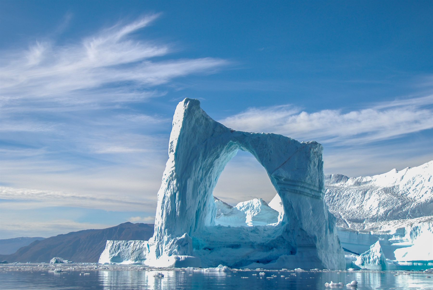 A towering iceberg with a natural arch in Arctic waters below snowy mountains